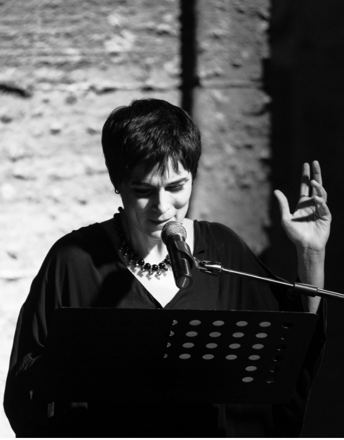 Lettura poetica dal vivo su palco con microfono e leggio, performance di poesia e musica in teatro, atmosfera intima in bianco e nero.